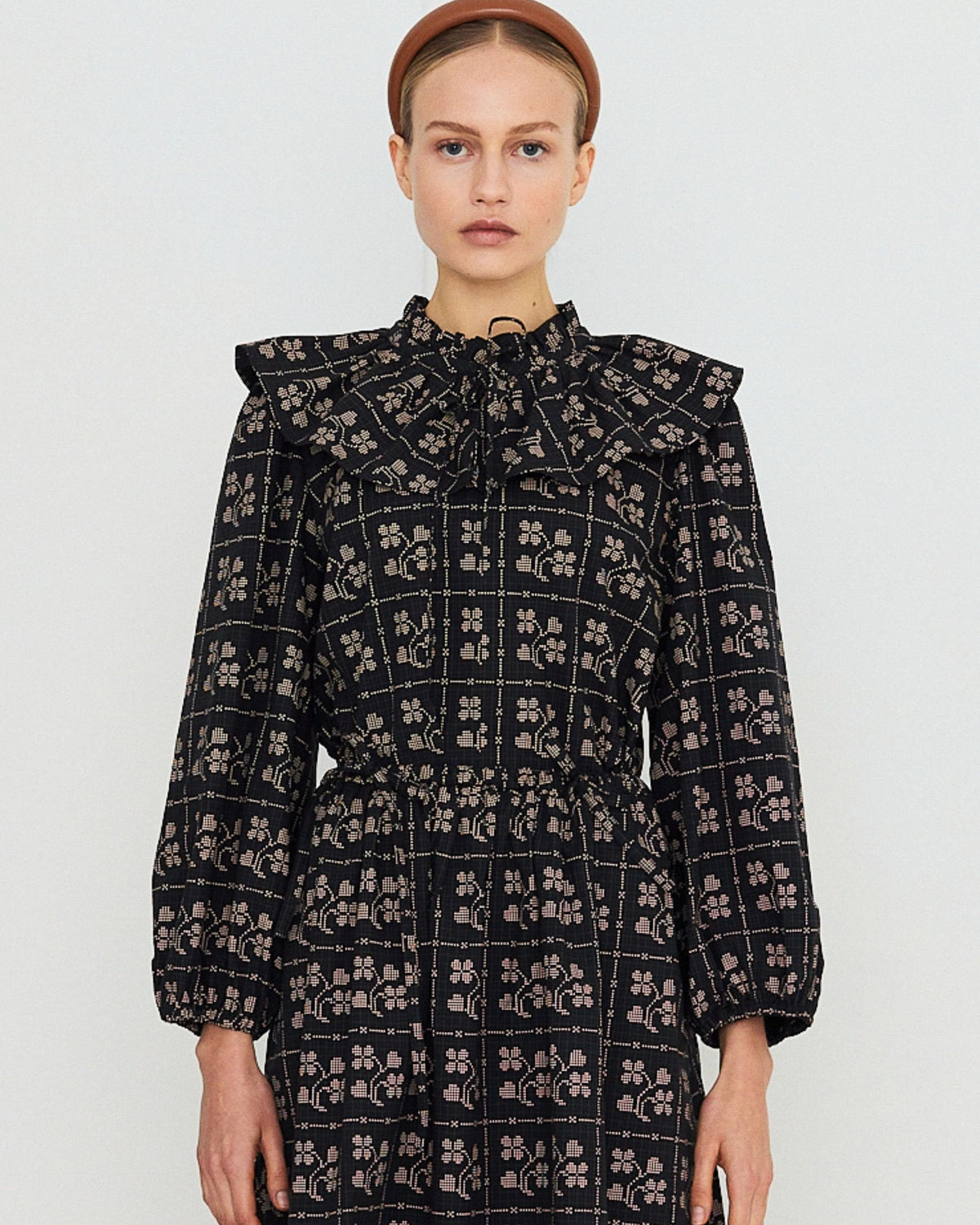 A model wearing a long-sleeved black dress with a beige floral grid pattern, featuring a high ruffled collar and a matching headband, set against a plain light gray background.